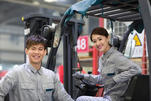 Confident Forklift Operators After UMW Training Two smiling forklift operators, a man and a woman, in a UMW Equipment warehouse after completing their training