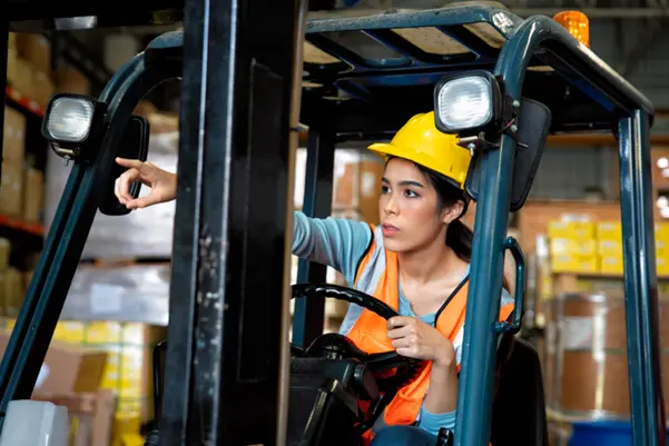 Woman Receiving Forklift Training A woman in a hard hat and safety vest operating a forklift in a warehouse setting