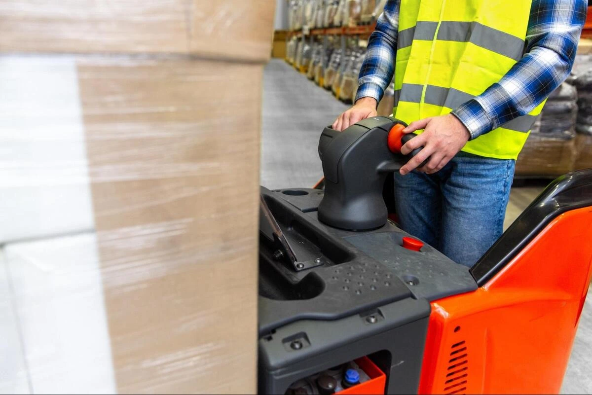 Operator using a powered pallet truck in a warehouse A warehouse worker operating an orange electric powered pallet truck to move boxes.