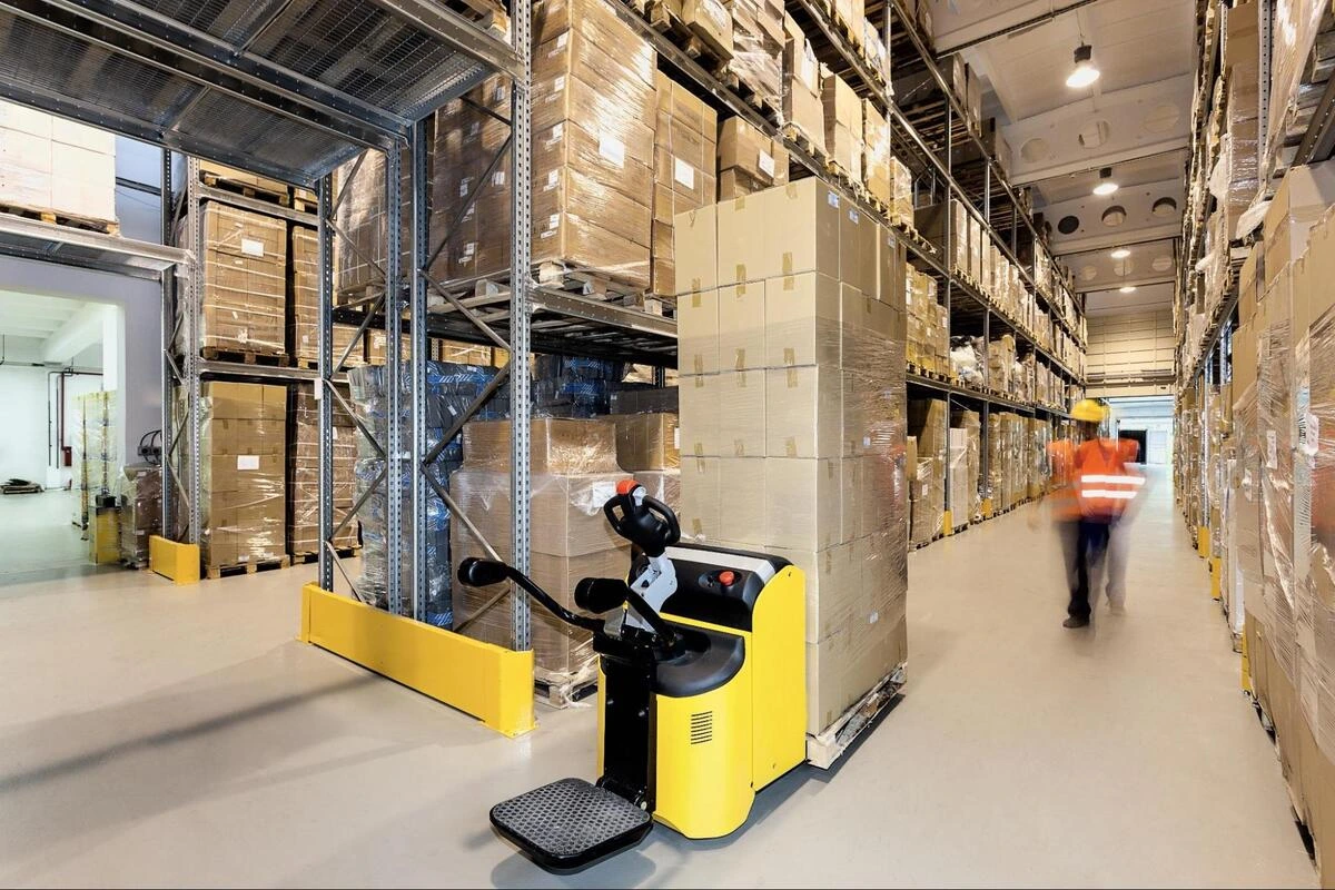 Electric pallet truck navigating a narrow warehouse aisle A yellow powered pallet truck moving through a narrow aisle in a high-rack warehouse facility