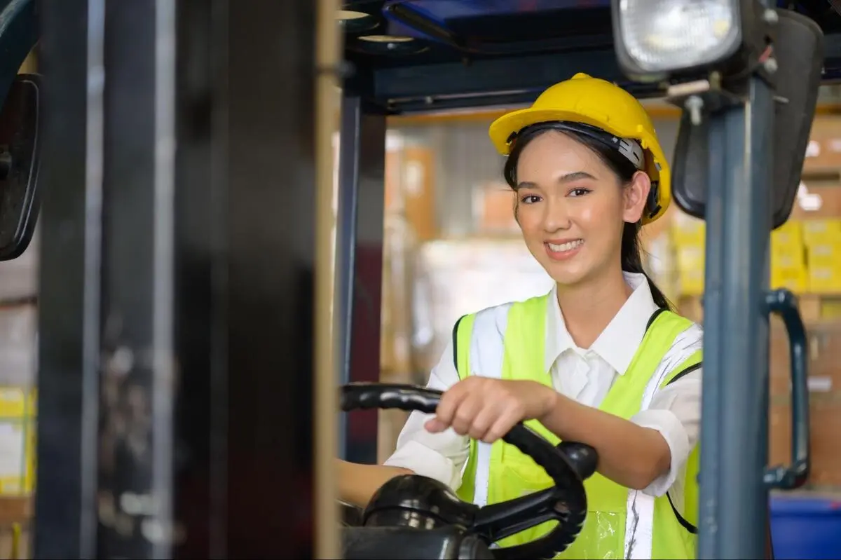 Female Forklift Operator in Warehouse Safety Gear Female warehouse worker in yellow vest and hard hat operating a forklift while moving goods in a warehouse