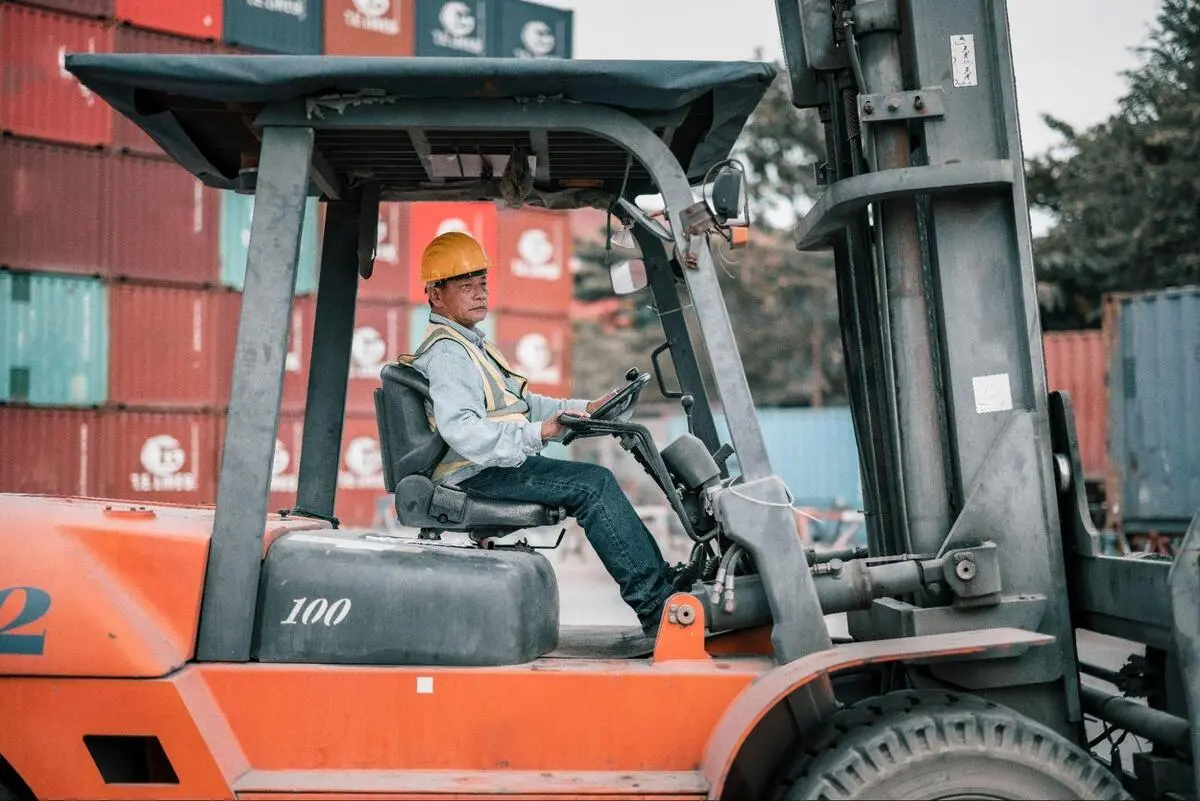 Industrial Forklift Operation in Shipping Yard A male operator driving a heavy-duty forklift near shipping containers, suitable for outdoor logistics and heavy load transport.