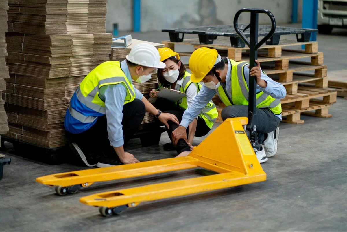 Powered Pallet Truck Safety Inspection Team Three warehouse workers inspecting a yellow powered pallet truck in a warehouse setting.